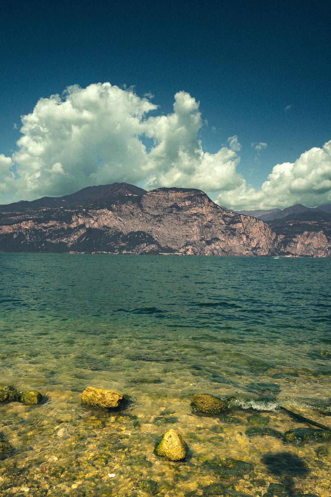 A tall cumulus cloud over the far ridge of Lake Garda, mossed stones resting in clear shallow water in the foreground.