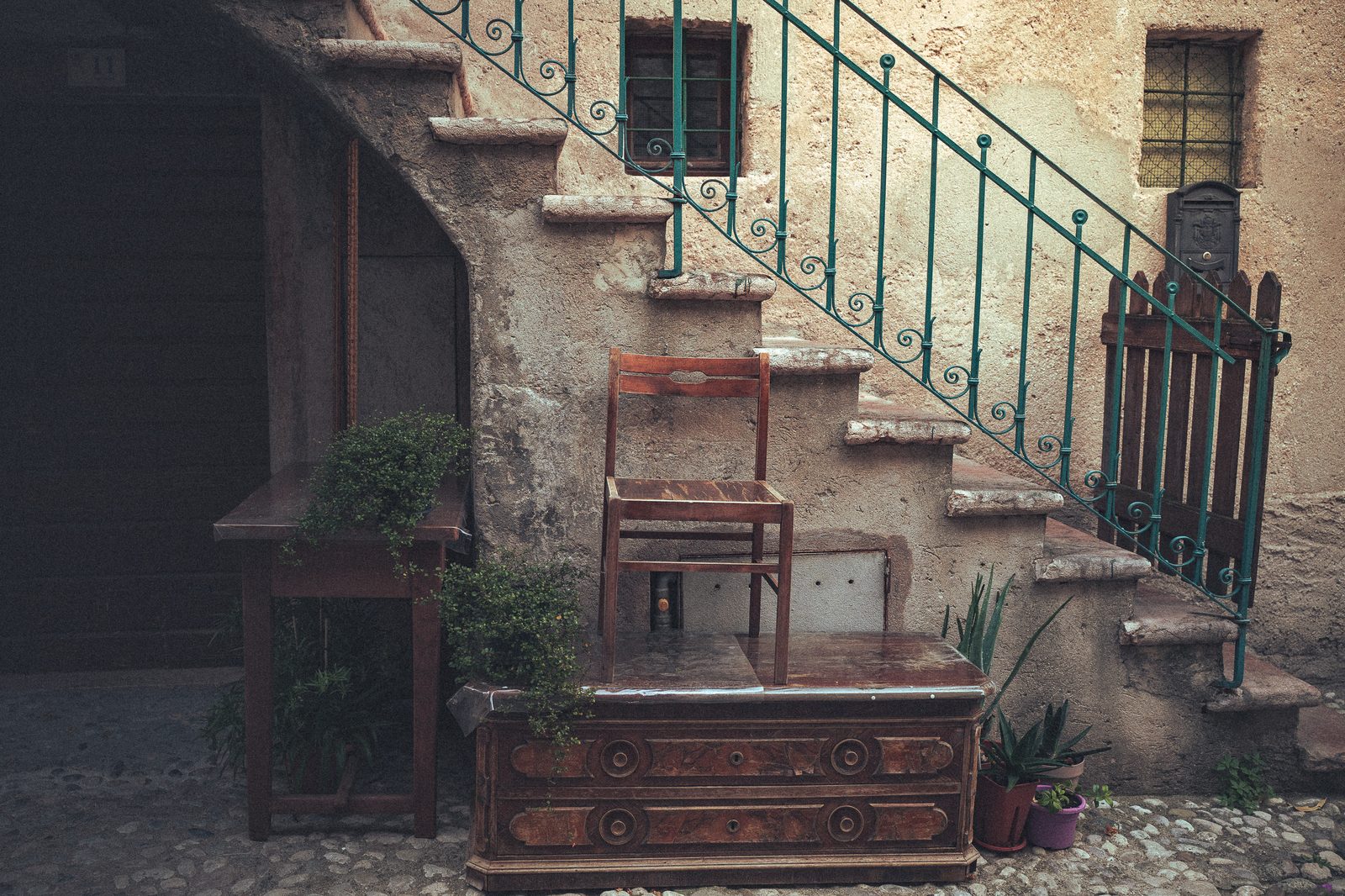 A wooden chair standing on a marble topped dresser in a stone courtyard, beside a green iron stair rail.