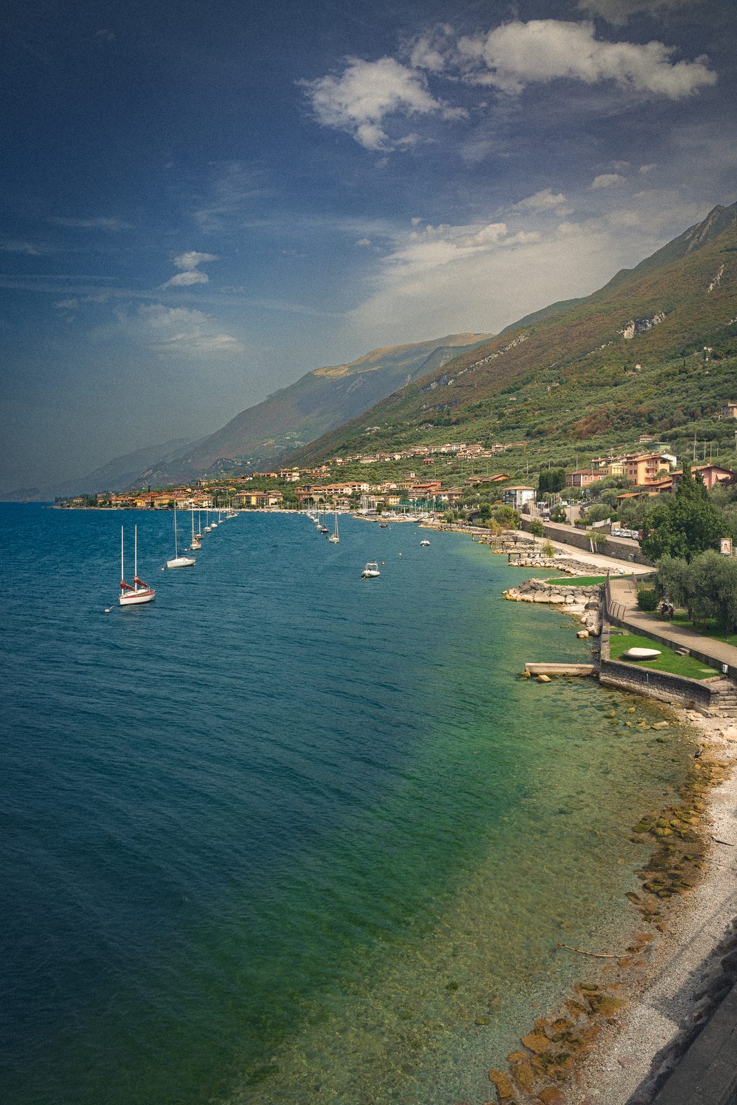 The eastern shore of Lake Garda from above, moored sailboats on clear green water, the town strung along the hillside.