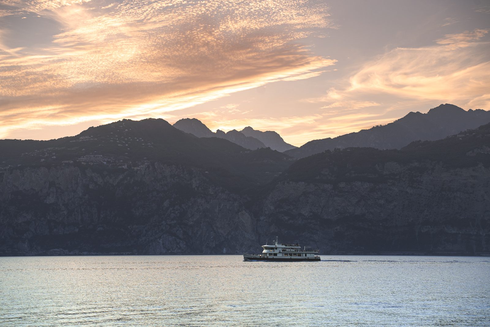 A passenger ferry crossing Lake Garda at sunset, silhouetted against the far cliffs under an orange sky.