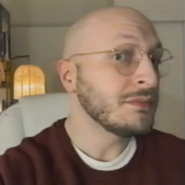 A bald man in glasses and a maroon sweater seated in a living room.