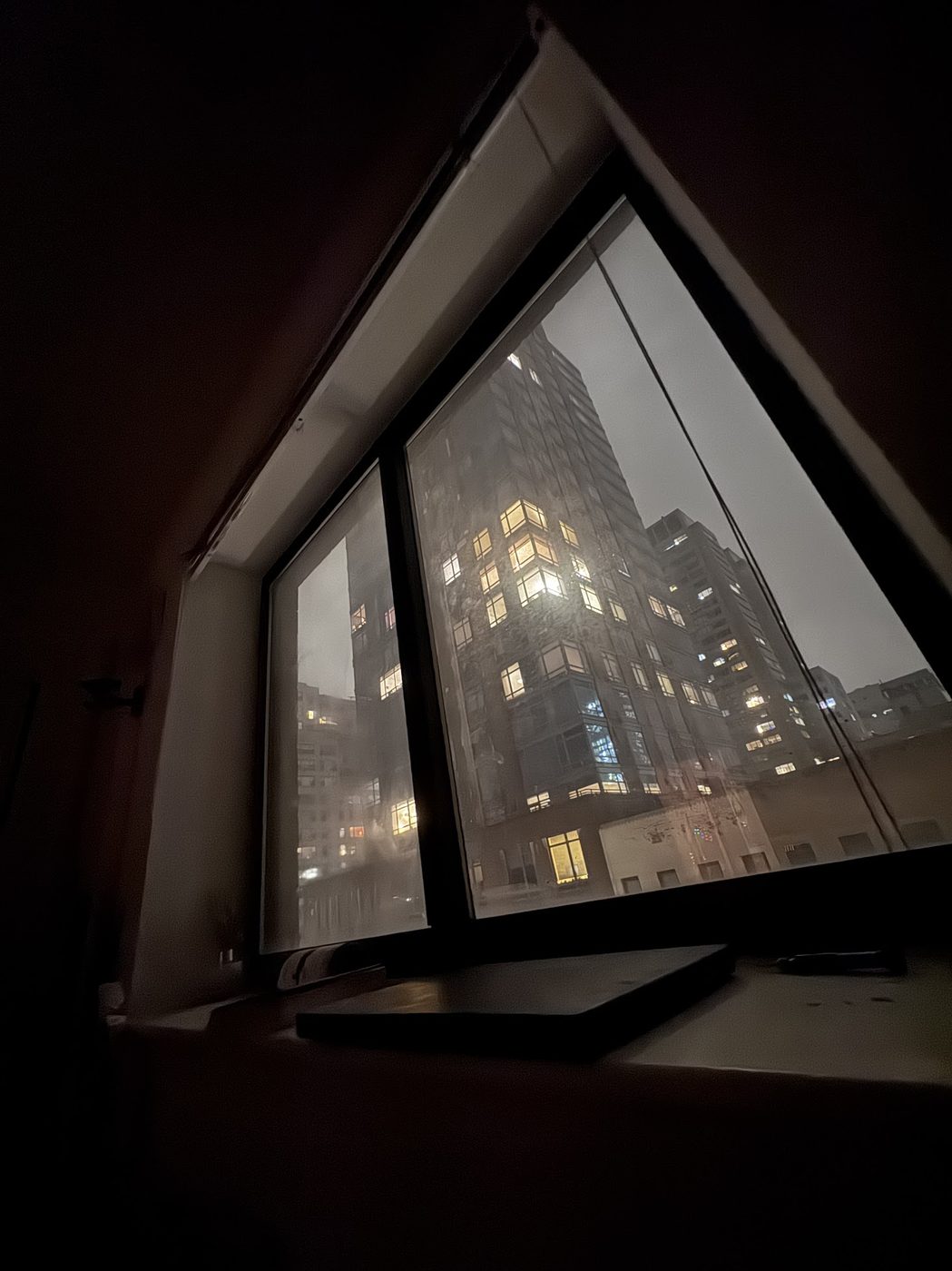 A dark room at night, an angled window above a desk, a residential tower with scattered lit windows outside.
